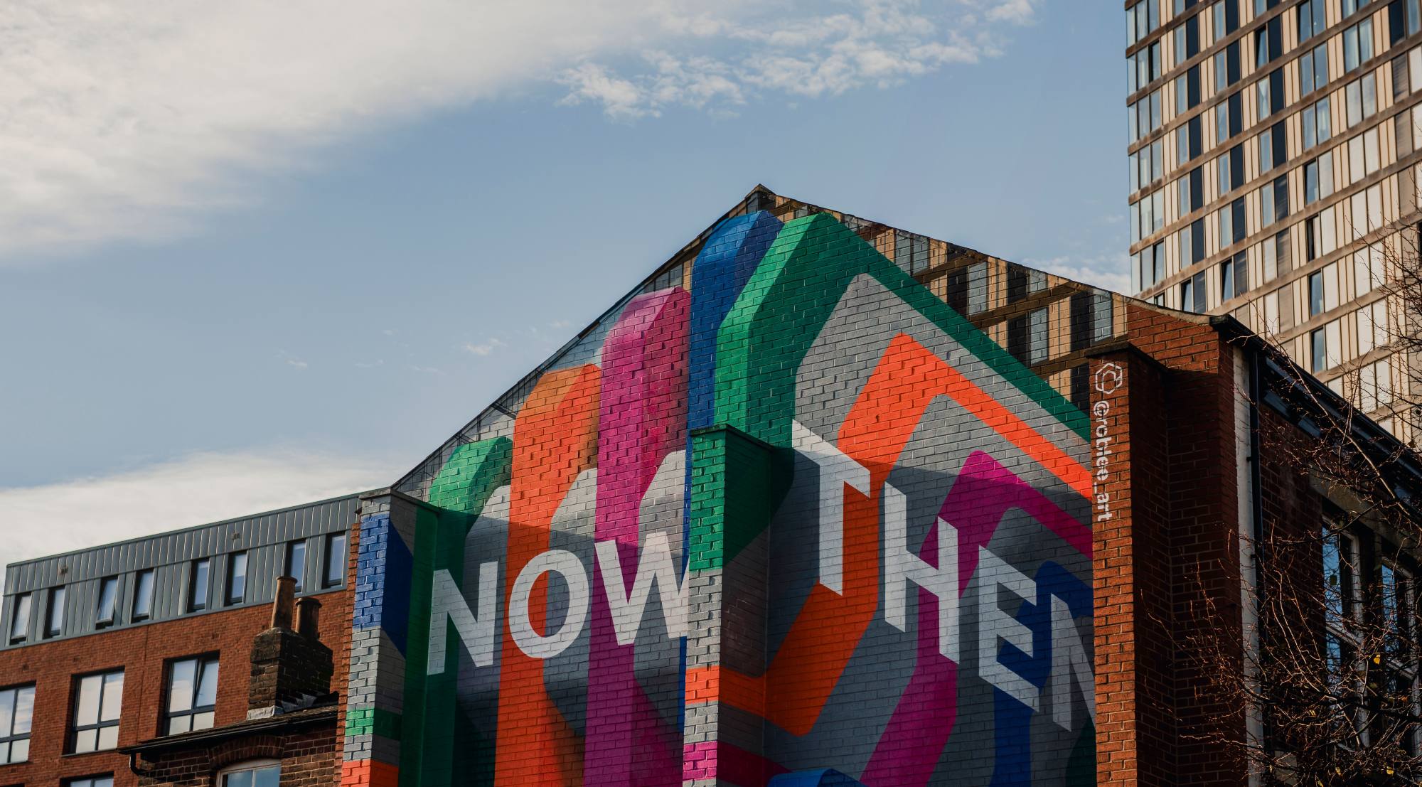 A colourful mural on a building wall with the words "NOW THEN" in large white letters, surrounded by geometric shapes in different colours. Modern urban flats are visible in the background.