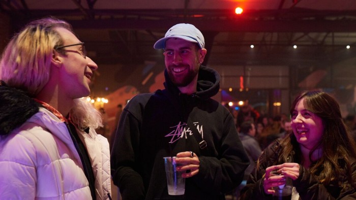 Three people standing together indoors talking happily, holding drinks in a dimly lit street food market.