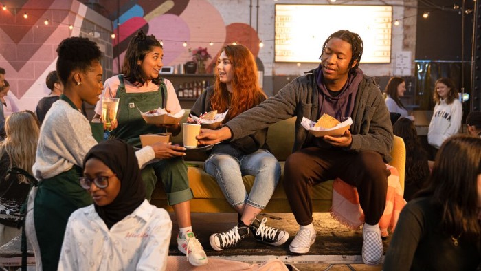Several young people sitting on a couch in a street food market setting with food containers, string lights, and colourful wall art.