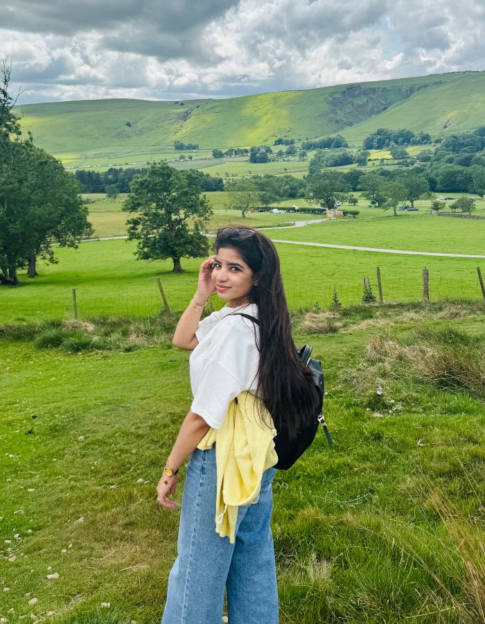 A young person standing in a grassy field in the Peak District, with hills, trees, and a partly cloudy sky in the background.