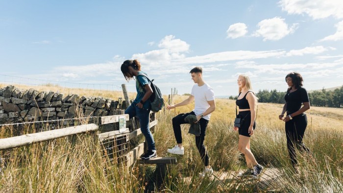 Four students hiking in a grassy field in the peak district, with one climbing over a wooden stile beside a stone wall under a sunny blue sky.