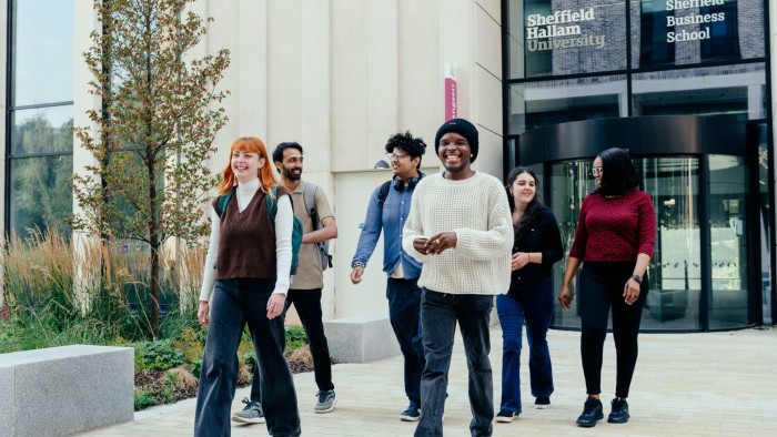 A group of people walk outside Sheffield Hallam University's Business School, with glass windows, a revolving door, and greenery on the left.