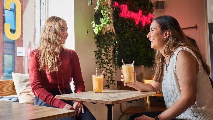 Two students sat in a cafe drinking juice, chatting and smiling. 