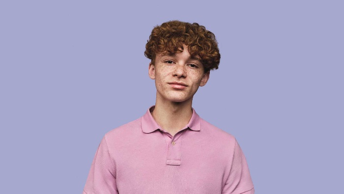 Against a purple background, a young man with curly hair wearing a pink polo shirt is smiling at the camera.