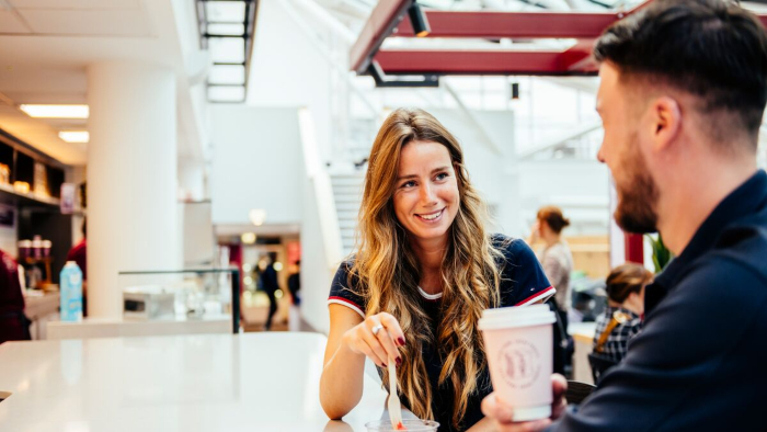 A man and woman are sat at a table in a cafe on campus, engaged in conversation over coffee.