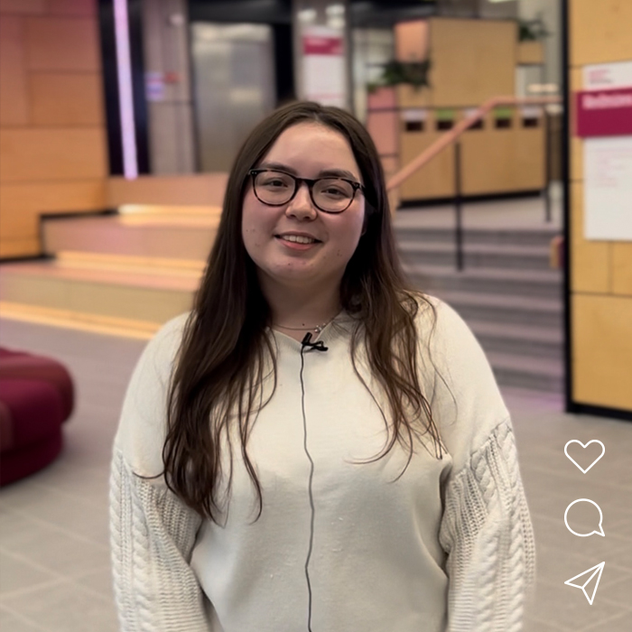 A woman in a white jumper wearing glasses is standing in a university building, smiling at the camera.