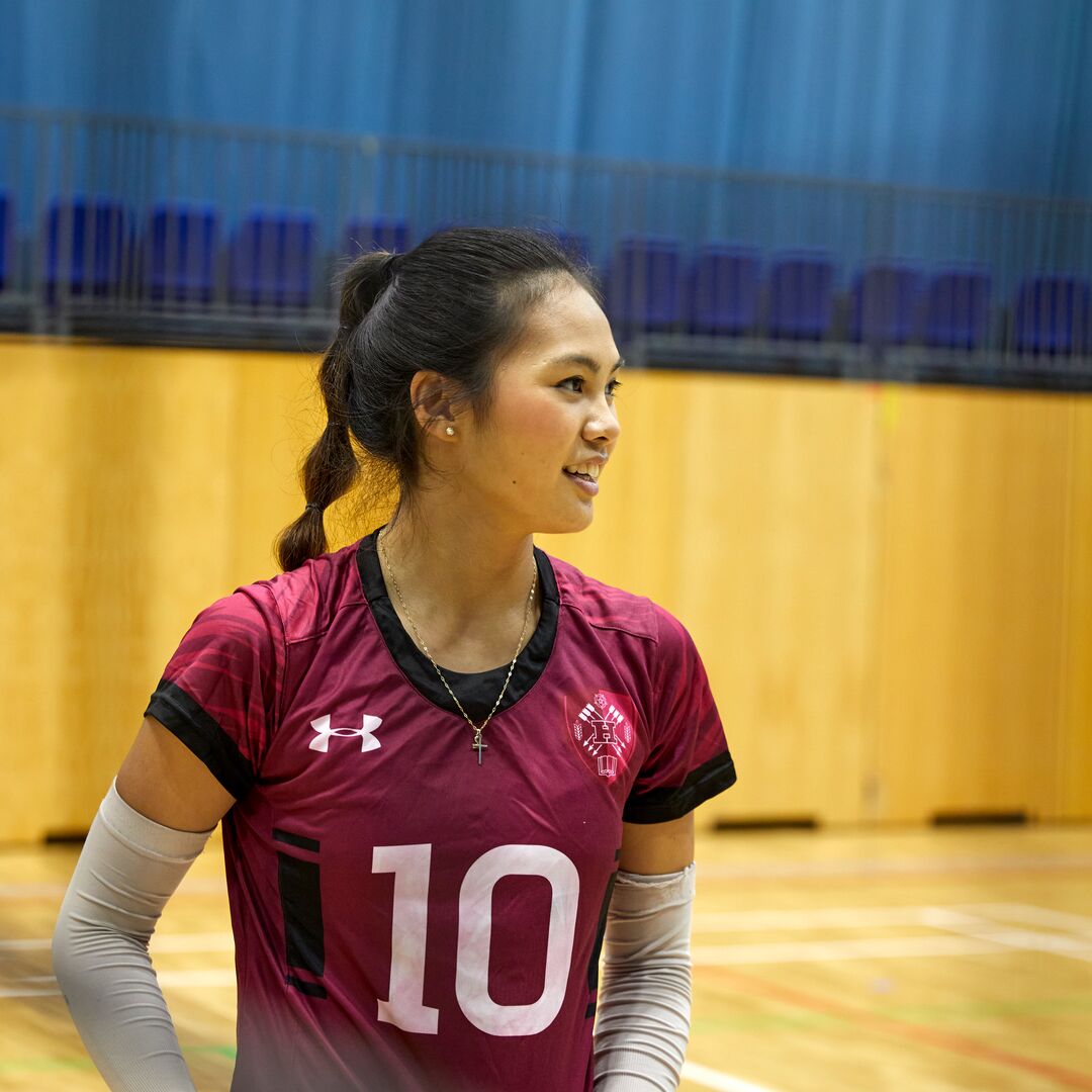 A young woman in a volleyball uniform stands on a court, smiling.