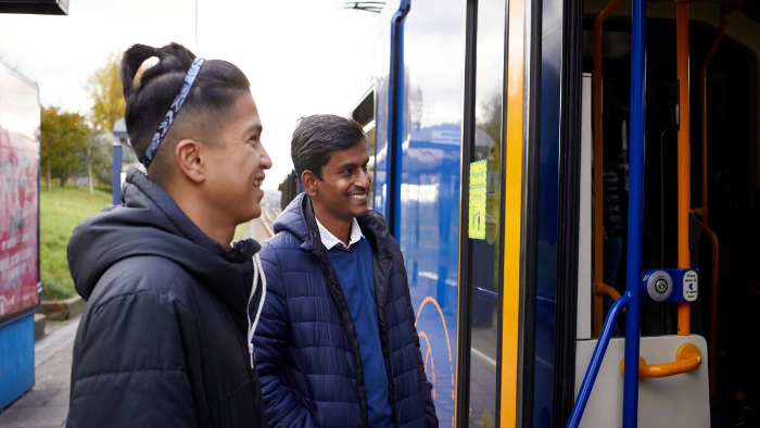Two young men stand beside a tram, smiling and engaged in conversation.