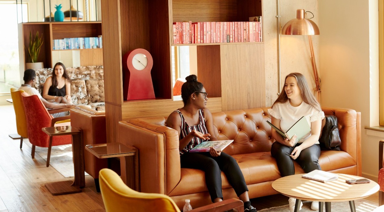 Two students sat on a sofa in their halls of residence communal area. They both have a book open and are in a discussion.