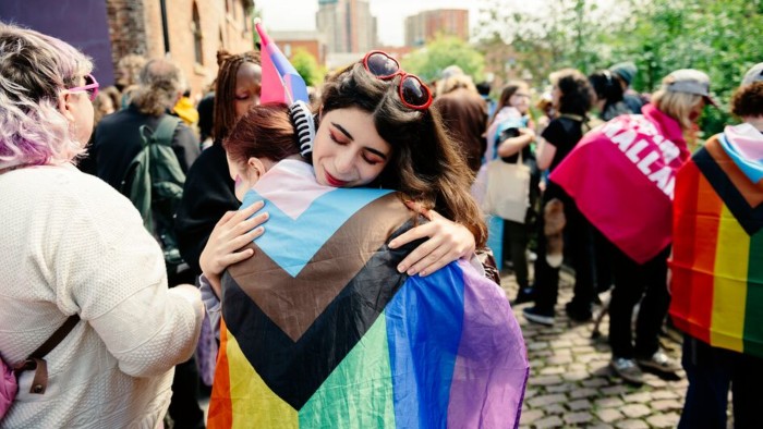 Two people hugging while wrapped in a rainbow Pride flag among a group of people at a Pride event.