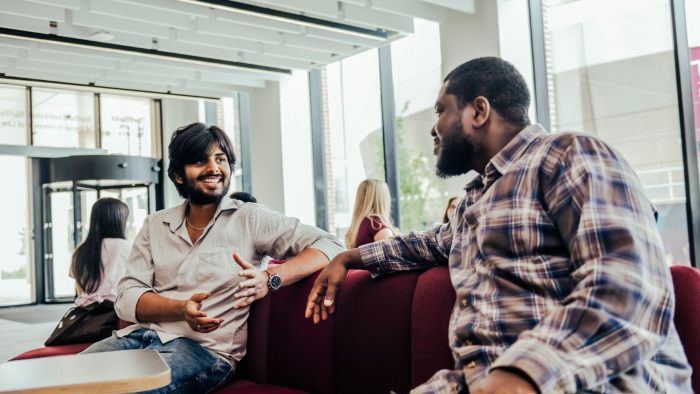 Two students sat in conversation on a large red sofa near a city campus building entrance. Two other students are sat nearby.