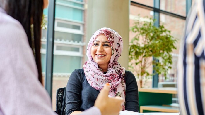 A student wearing a patterned headscarf is seated indoors at a table in a modern office, engaged in conversation with two others.