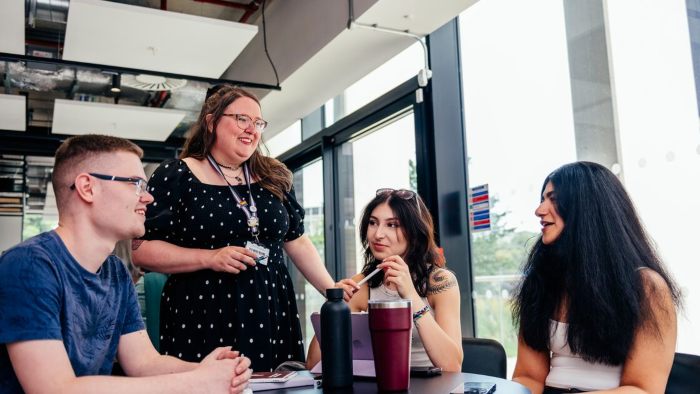 Three students sitting at a table at city campus with notebooks, cups, and bottles, while another person stands and talks to them.
