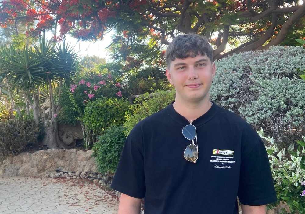 Connor is stood outside and wearing a dark blue t-shirt with sunglasses hooked over his collar, with Mediterranean plants and greenery in the background.