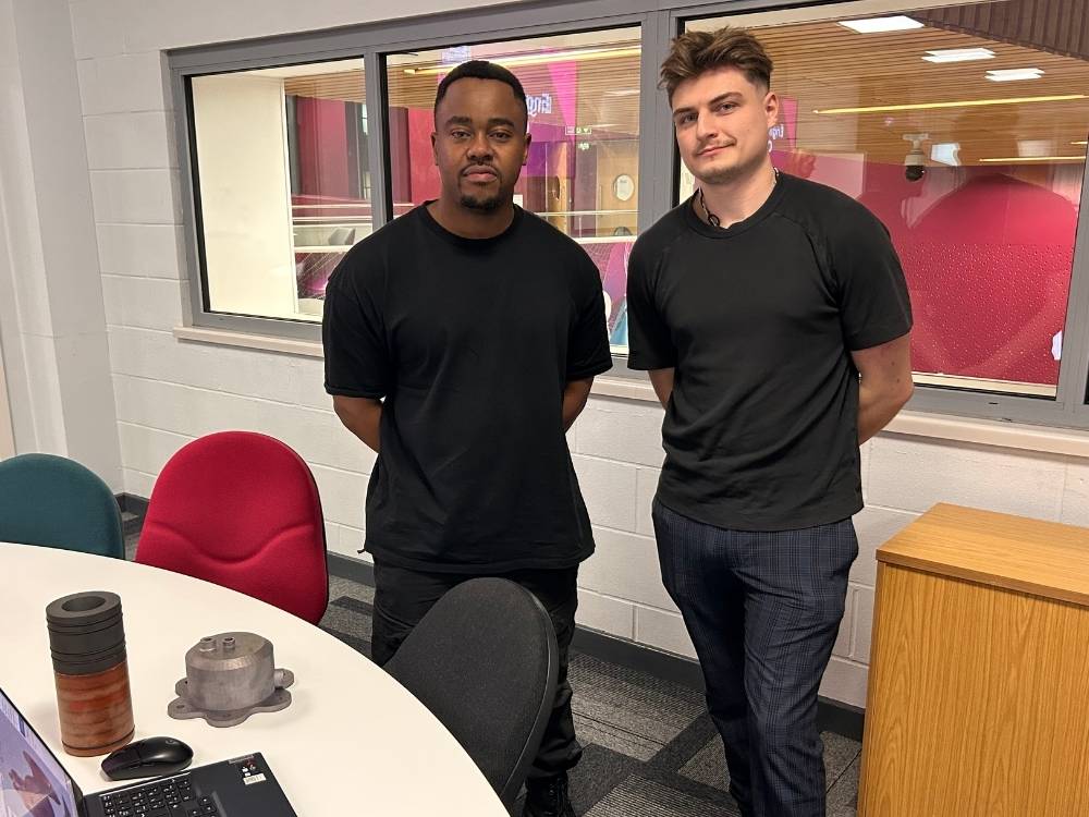 Two engineering students are stood together in a brightly lit room, both wearing black t-shirts, and there are two cylindrical metal components placed on a table in front of them.