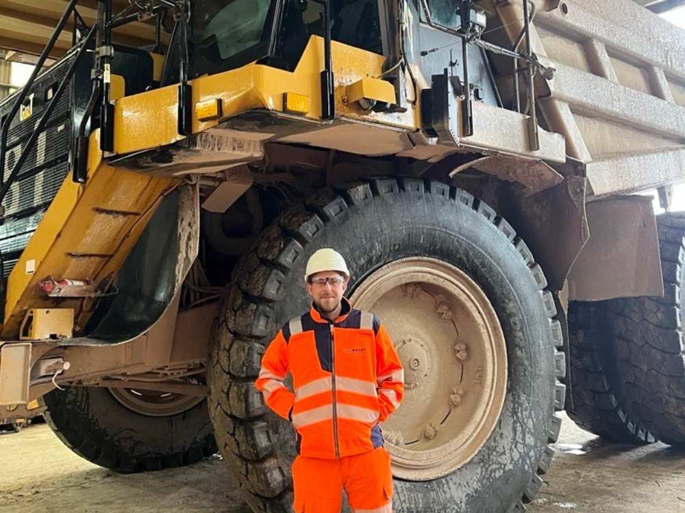Jacob is wearing orange hi-vis overalls and a white hard hat, stood in front of a large yellow construction site vehicle.