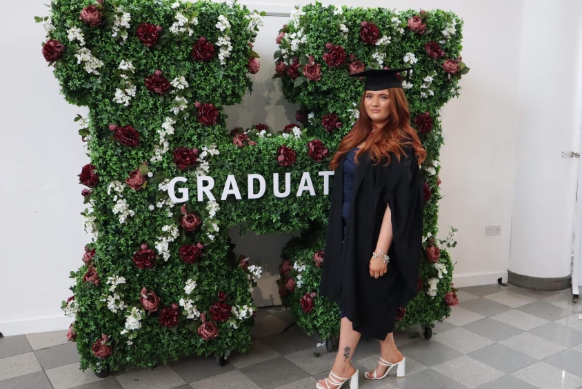 Jade Blackburn on graduation day. She is wearing a cap and gown and is standing in front of the big Hallam 'H'