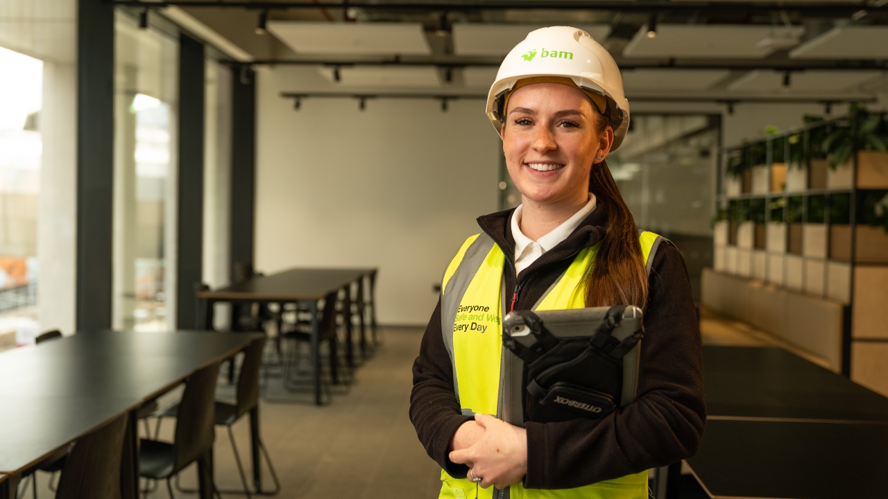 Jasmine Lidster is wearing a hard hat and a hi vis. She is standing on site in one of the University's new buildings. She is smiling and holding an ipad.