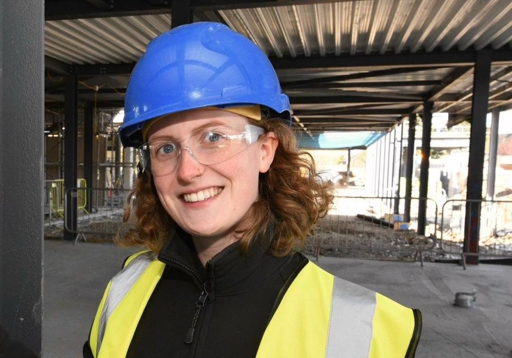 Katy is smiling, wearing a blue protective hard hat and hi-vis vest, stood in a partially constructed building.