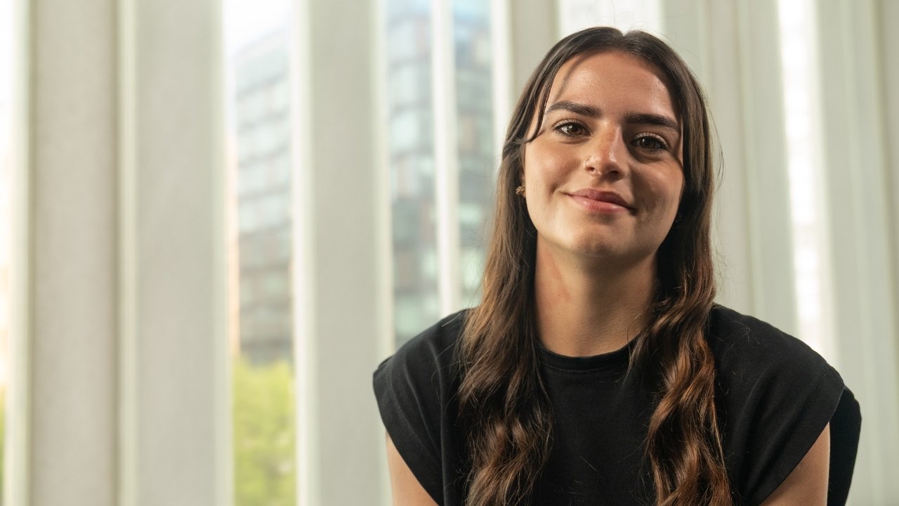 Leah is smiling and wearing a black t-shirt, with windows in the background showing a view of trees and a tall city building.