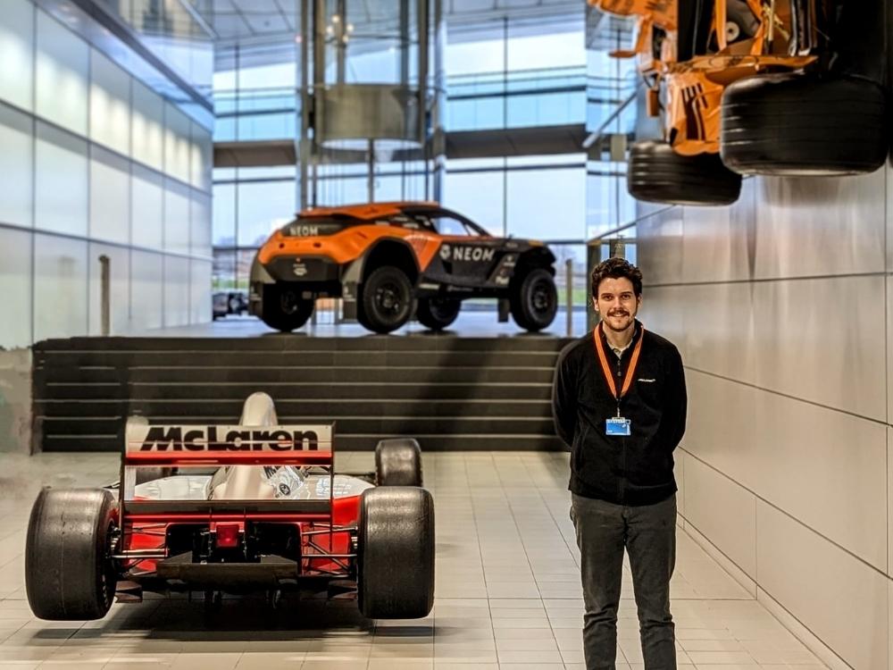 Nathan is wearing a black fleece and orange lanyard, stood next to an orange and white race car with the McLaren logo displayed on it's spoiler. There are two other race cars in the background.