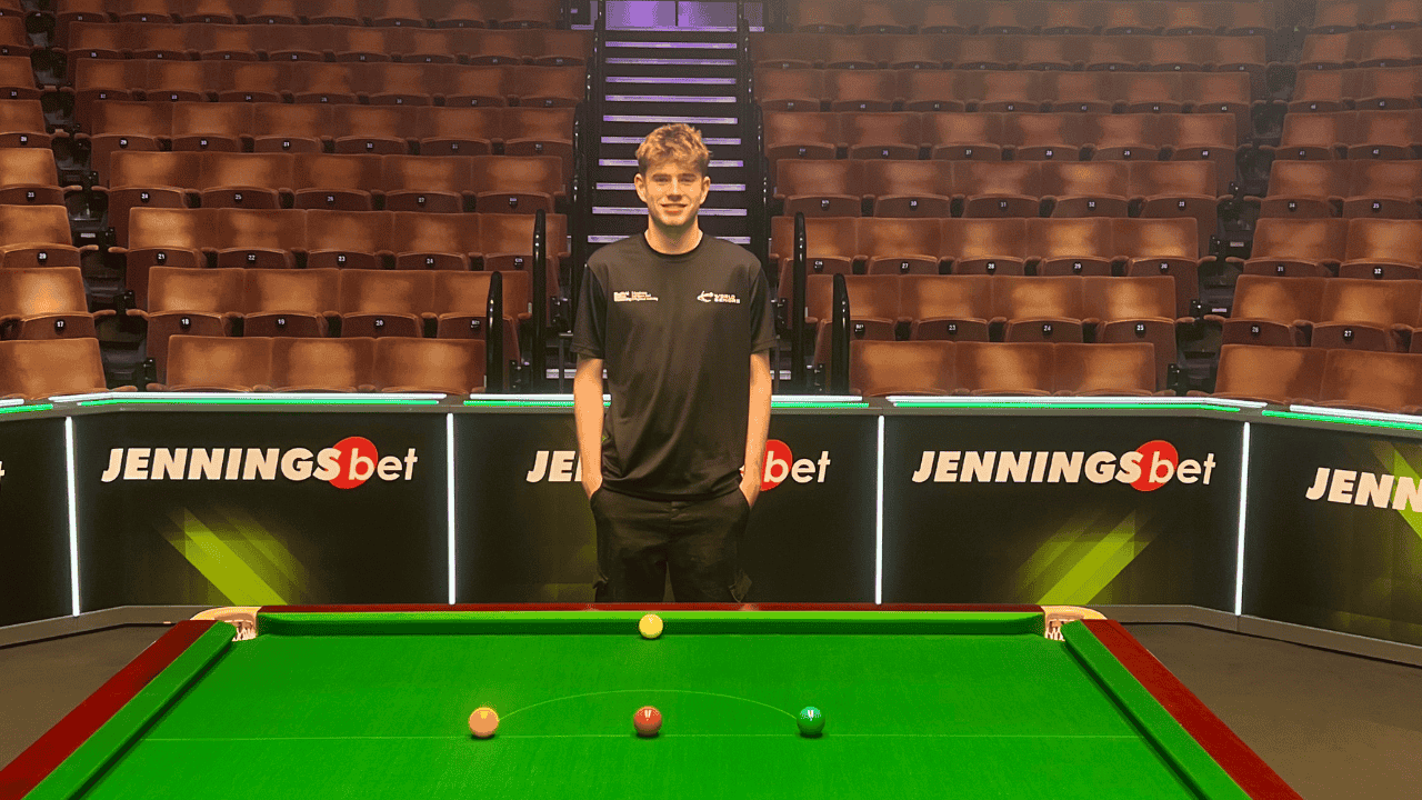 Nathan is wearing a black t-shirt with a white World Snooker logo and is stood behind a snooker table, with tiered rows of orange seats behind him.