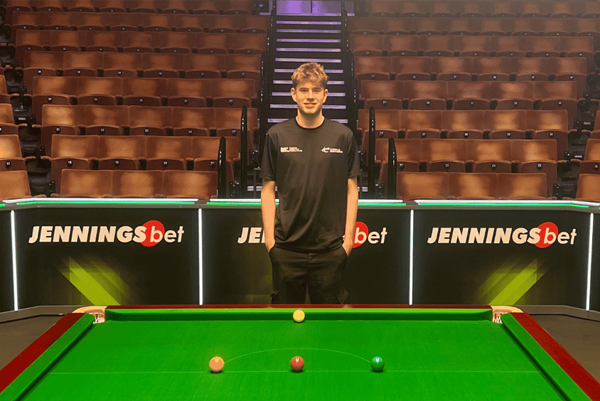 Nathan is wearing a black t-shirt with a white World Snooker logo and is stood behind a snooker table, with tiered rows of orange seats behind him.