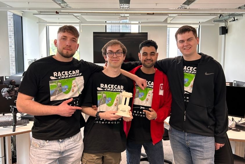 Four male students are stood together and wearing black t-shirts with the words 'Racecar Crashers' printed on. The student in the middle is holding a white trophy.