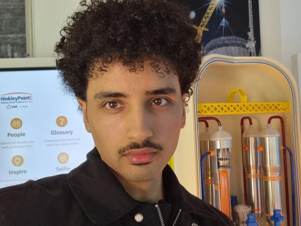 Head-and-shoulders photo of Shehab, who is wearing a black jacket. There is a 3D model of silver energy cylinders and a computer screen behind him.