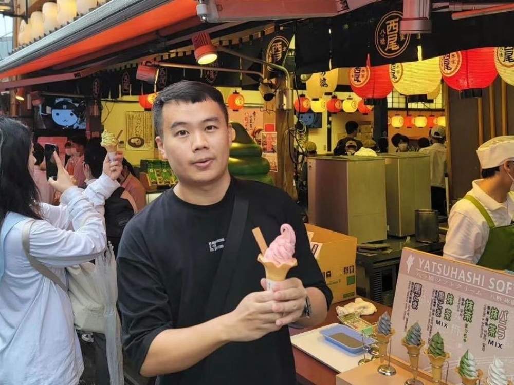 Zhenfeng Lu is wearing a black t-shirt and holding an ice cream cone, stood in a busy street with market stalls and hanging-lanterns to his right.