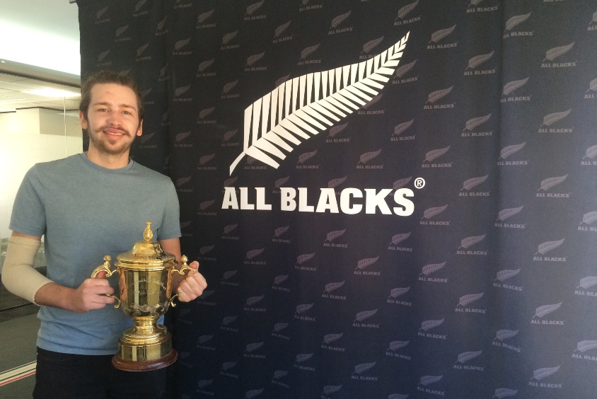 White male in front of New Zealand All Blacks sign holding a trophy.