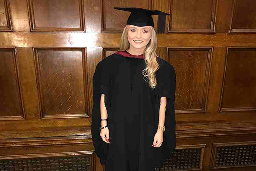 Blonde girl wearing smiling at the camera wearing a graduation gown and cap.