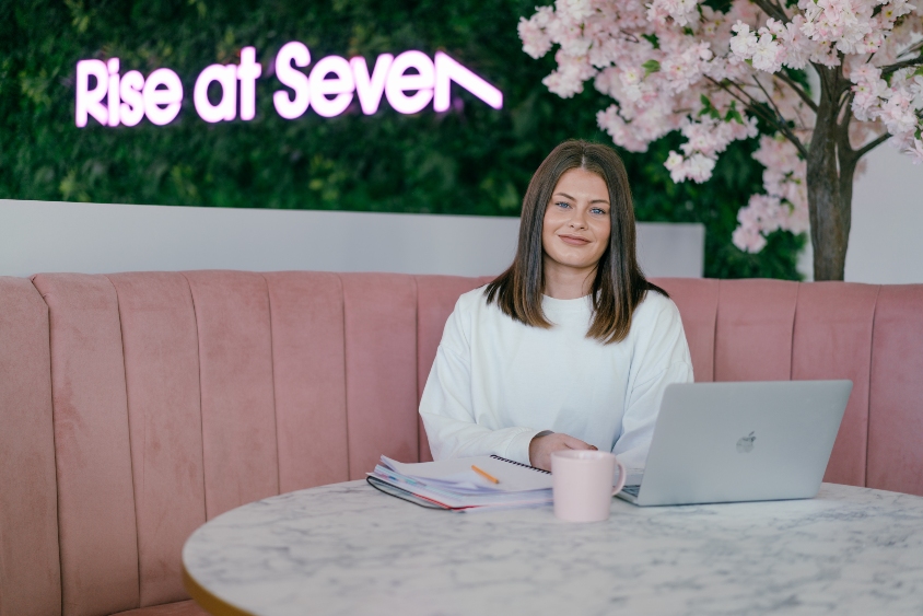 Woman with brown hair and white jumper on a pink booth seat with a laptop, notepad and mug in front of her on a table.