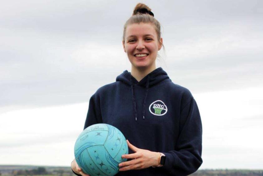 Izzy stands outside, smiling and holding a light blue netball. She wears a dark blue hoodie. 