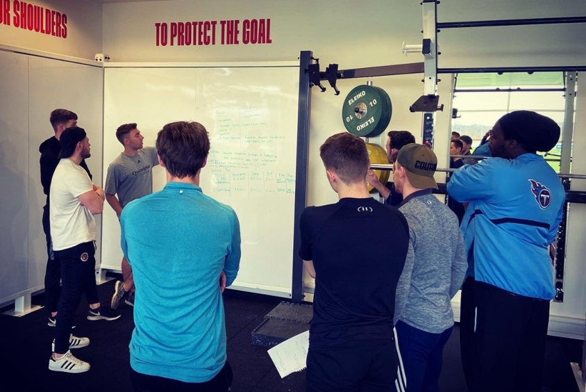 A group of students stood up gathered around a floor length whiteboard in the sports facilities with Matt pointing at the board.