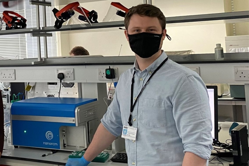 Male wearing blue shirt, protective gloves and face mask stands in a science lab with equipment on shelves behind them. 