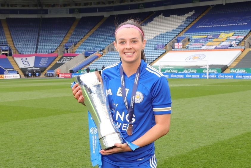 Female brunette wearing blue football kit stands centre of frame on the pitch at Leicester City's King Power Stadium smiling as she proudly cradles a Women's Championship trophy whilst also wearing a Women's Championship medal