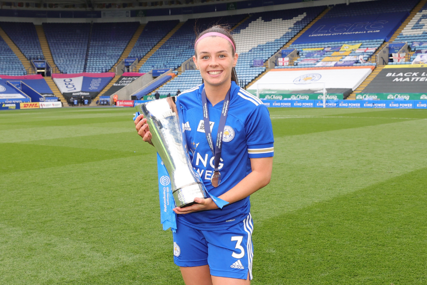 Female brunette wearing blue football kit stands centre of frame on the pitch at Leicester City's King Power Stadium smiling as she proudly cradles a Women's Championship trophy whilst also wearing a Women's Championship medal
