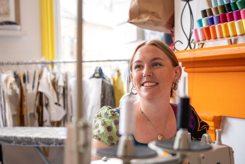 Image of long haired female designer sat at a sewing machine table with rail of garments behind her.