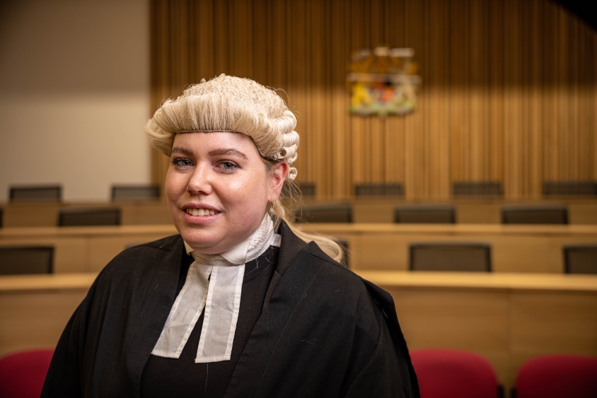 Female law student in legal barrister wig and gown stood in a criminal moot courtoom with seats in the background.