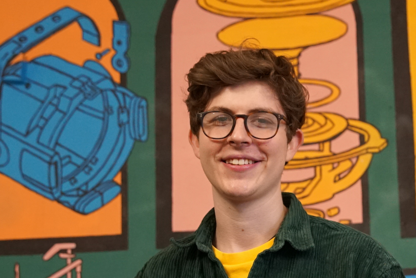 Male with glasses standing in front of a colourful mural smiling at the camera.