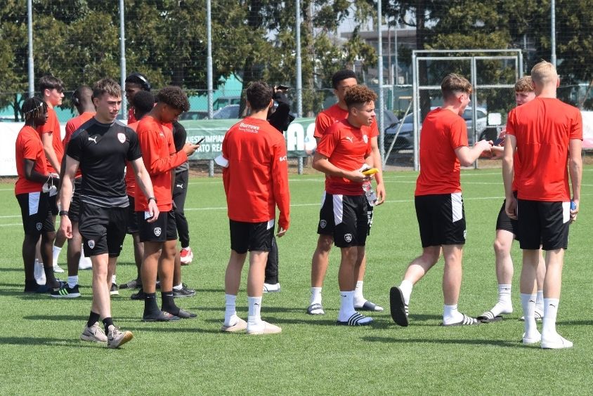 Shot haired male wearing a black football top and shorts walks in front of a group of younger males wearing orange coloured football kits.