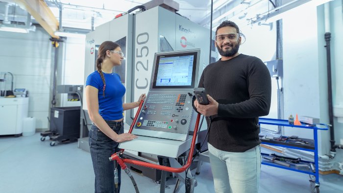 Two people wearing safety goggles in front of a large CNC machine—one using the control panel, the other holding a control device and smiling for a photo.