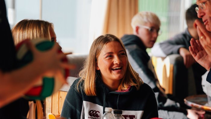 A group of people sitting indoors; one person in front wears a black and white hoodie and is smiling at a person talking off camera.