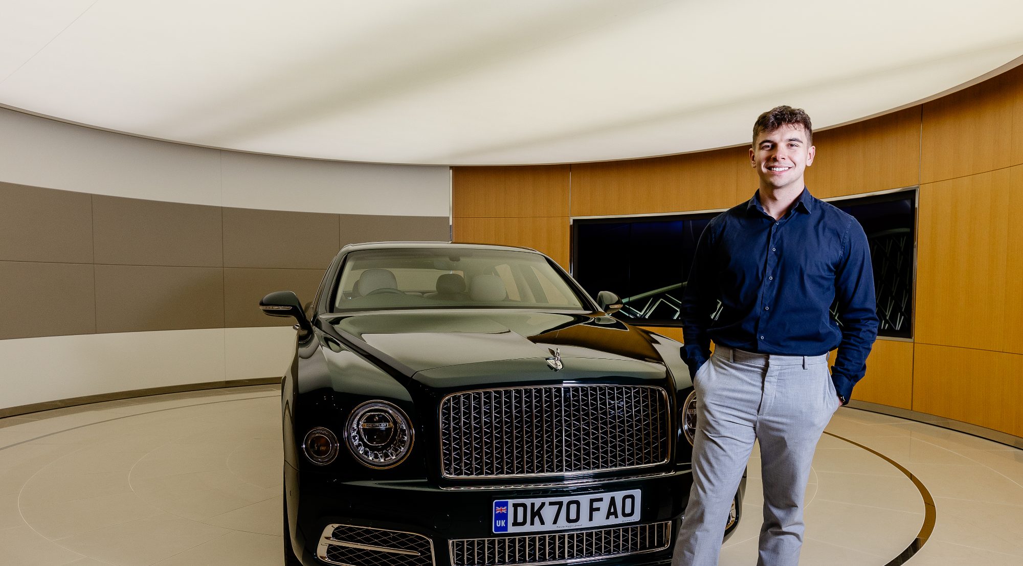 A man in a dark blue shirt and light grey trousers stands beside a black luxury car in an indoor showroom with curved walls.
