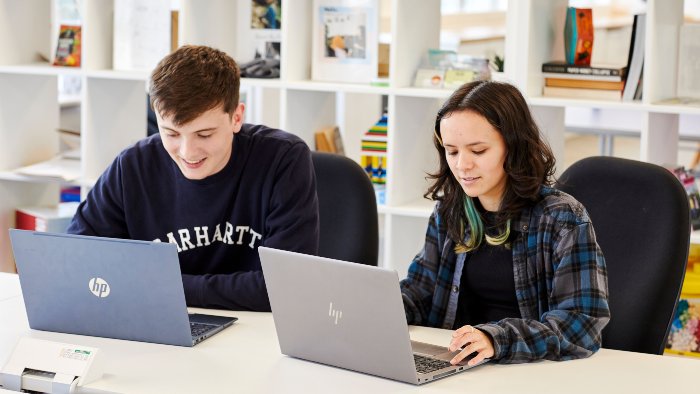  Two casually dressed people sit at a table in a well-lit communal room using laptops. Behind them, white shelves hold books, decorations, and office supplies.