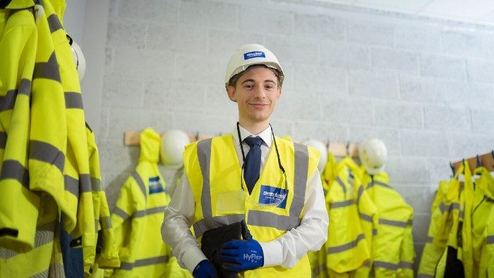 A Sheffield Hallam placement student at a construction firm. He is stood wearing a high visibility jacket, safety helmet and smiling for the camera.