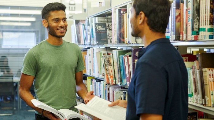 Two students stood talking to each other in a library at Sheffield Hallam. Both of them have a book open.