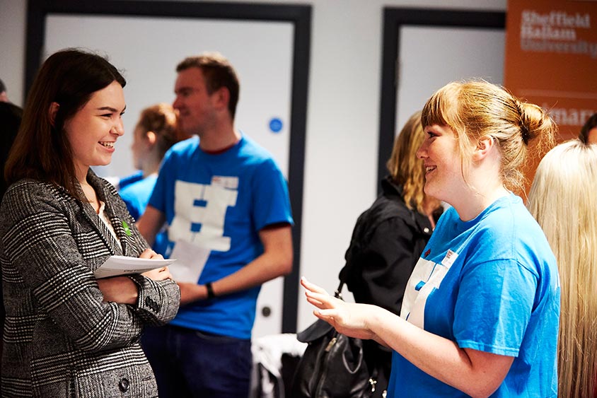 Two people talking at a Sheffield Hallam open day event. One holding a notepad, the other in a blue Team Hallam ambassador T-shirt.