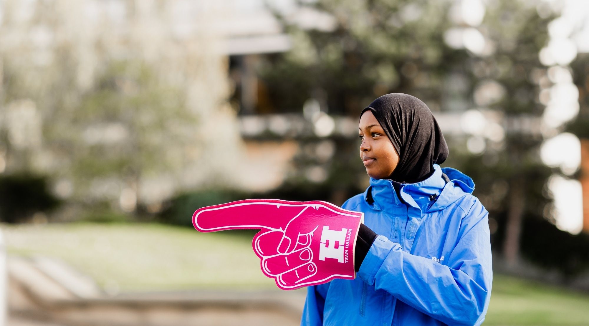 A young woman wearing a blue jacket stands outdoors, holding a large pink foam finger that points to the left. The foam finger has "TEAM HALLAM" printed on it.